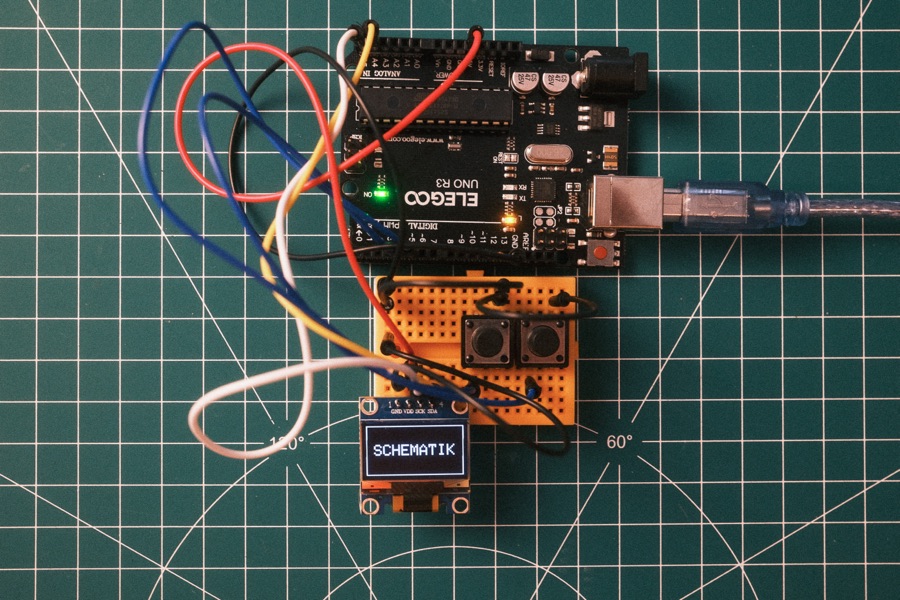 An Arduino board wired to a breadboard, two buttons and a small OLED display reading SCHEMATIK, on a cutting mat.